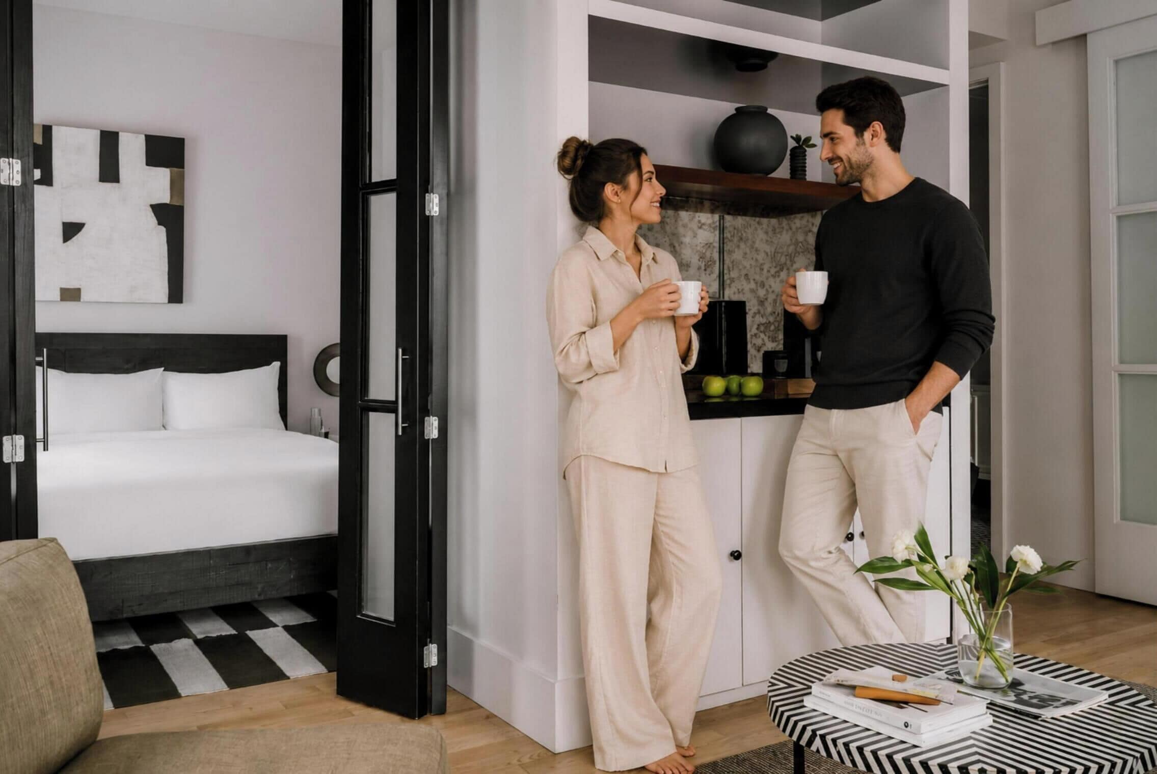 Un dormitorio moderno con cama, una mesita de noche con un libro y un jarrón, y una zona de cocina con frutero y planta en maceta.