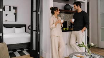 Un dormitorio moderno con cama, una mesita de noche con un libro y un jarrón, y una zona de cocina con frutero y planta en maceta.