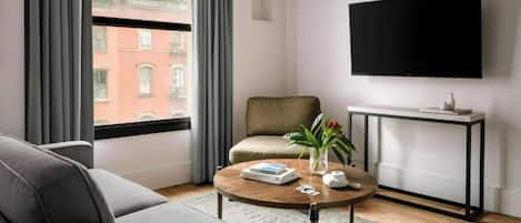 A modern living room with a sofa, a coffee table with books and a vase, a television mounted on the wall, and a view of a red brick building through large windows.