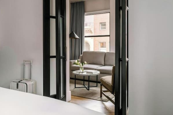 Moderni hotellihuone, jossa on sohva, pieni pyöreä pöytä maljakolla ja matkalaukku.