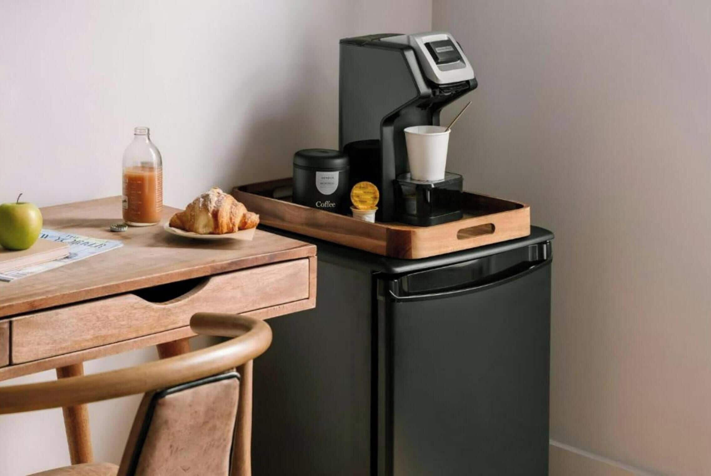 A kitchenette with a coffee machine, a croissant, a bottle of juice, and a green apple on a wooden table.