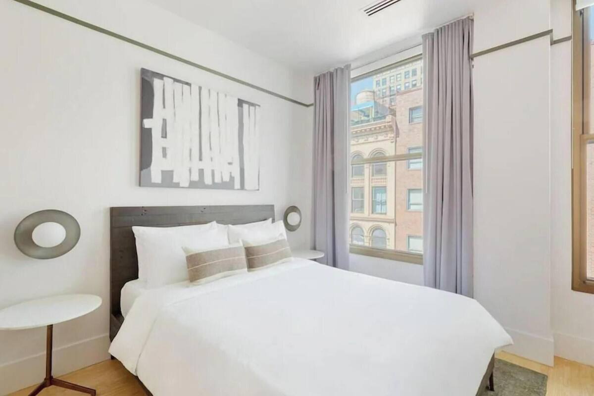 Une chambre d’hôtel avec un grand lit, une petite table ronde, une œuvre d’art accrochée au mur et une fenêtre avec des rideaux.