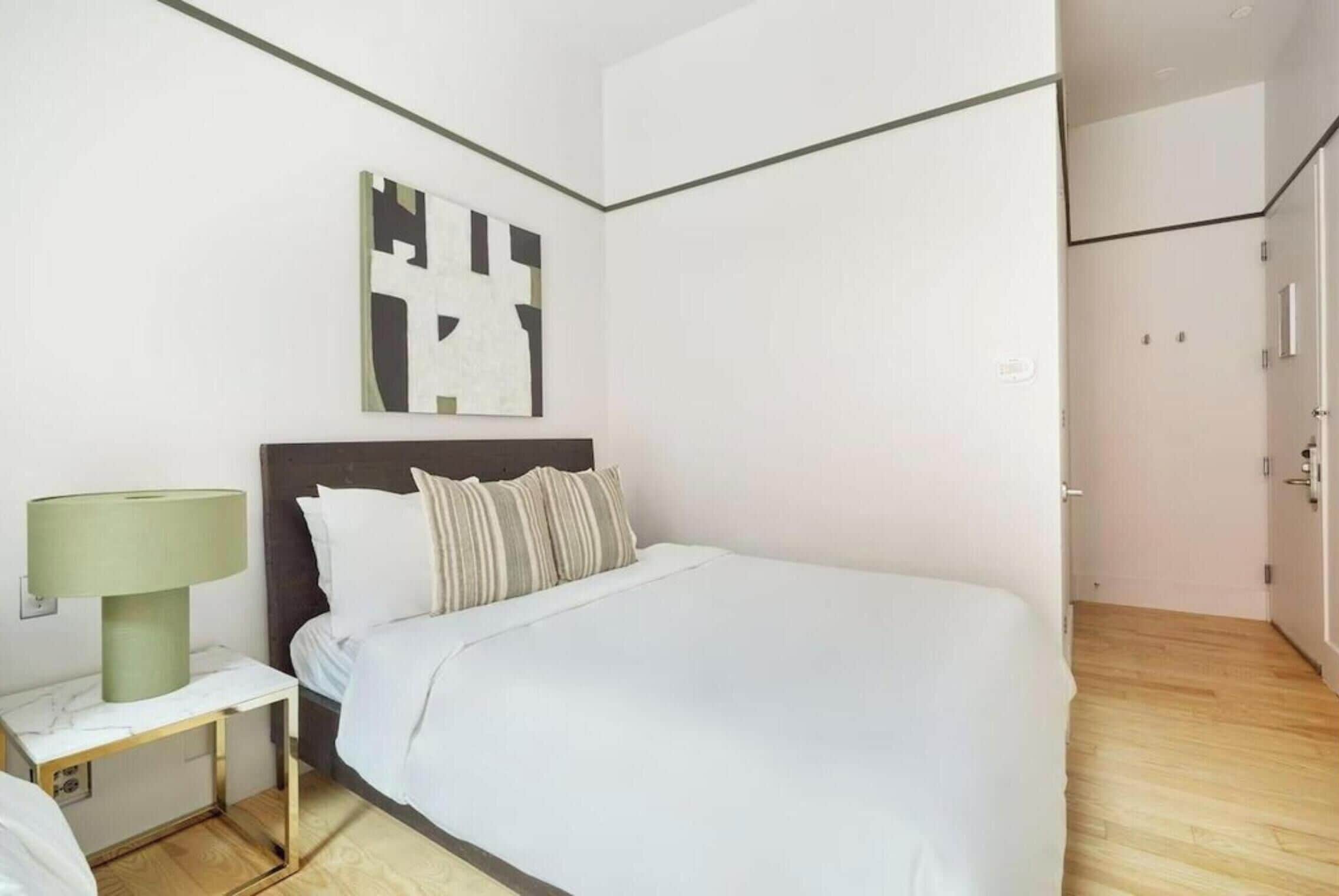 A modern hotel room with a large bed, a nightstand with a lamp, and a minimalist wall art piece.
