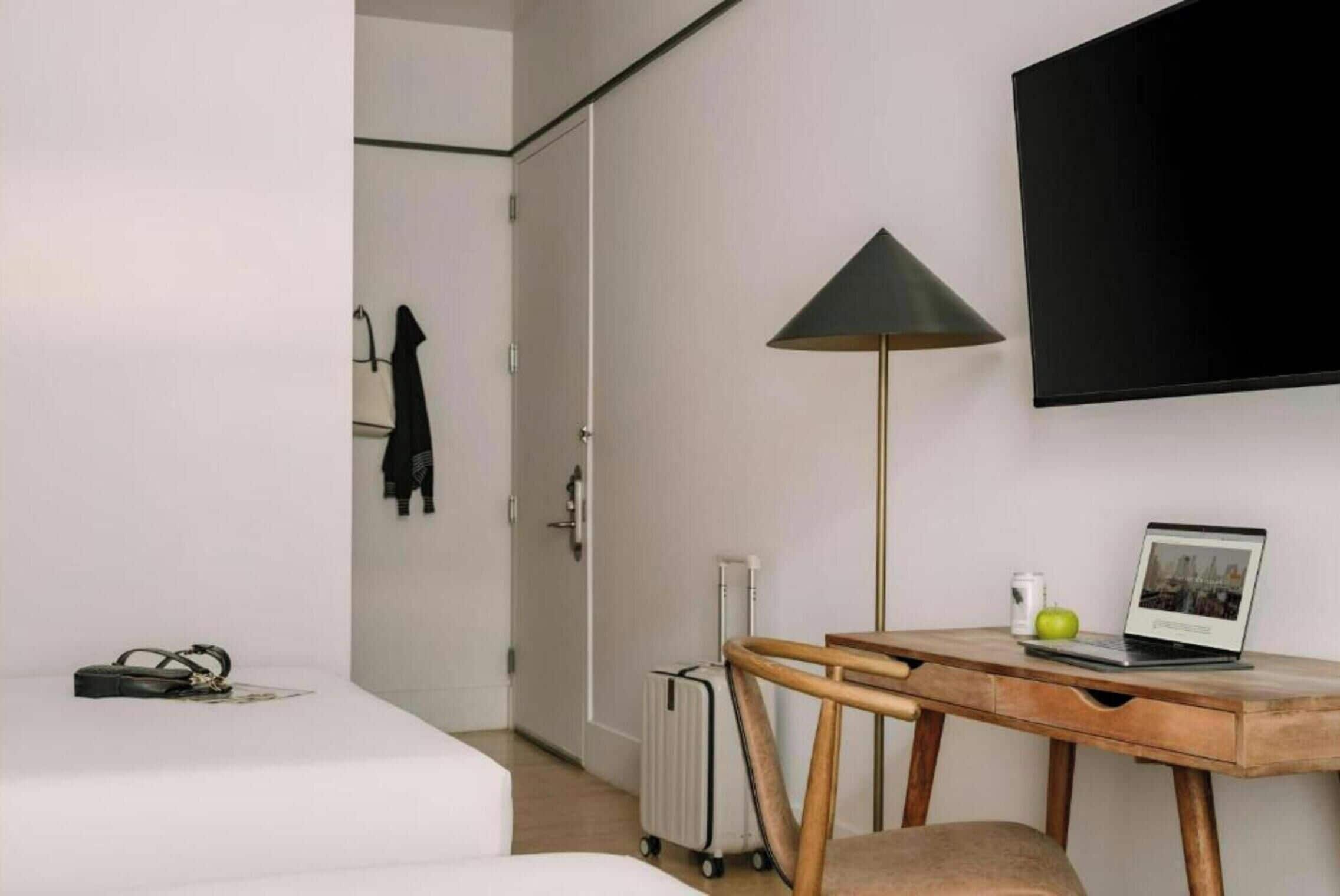 A hotel room with a bed, a desk with a laptop, a standing lamp, a radiator, and a coat rack.