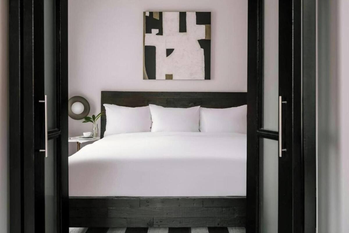 Un lit avec du linge de lit blanc, une tête de lit sombre et une œuvre d’art abstraite accrochée au mur.