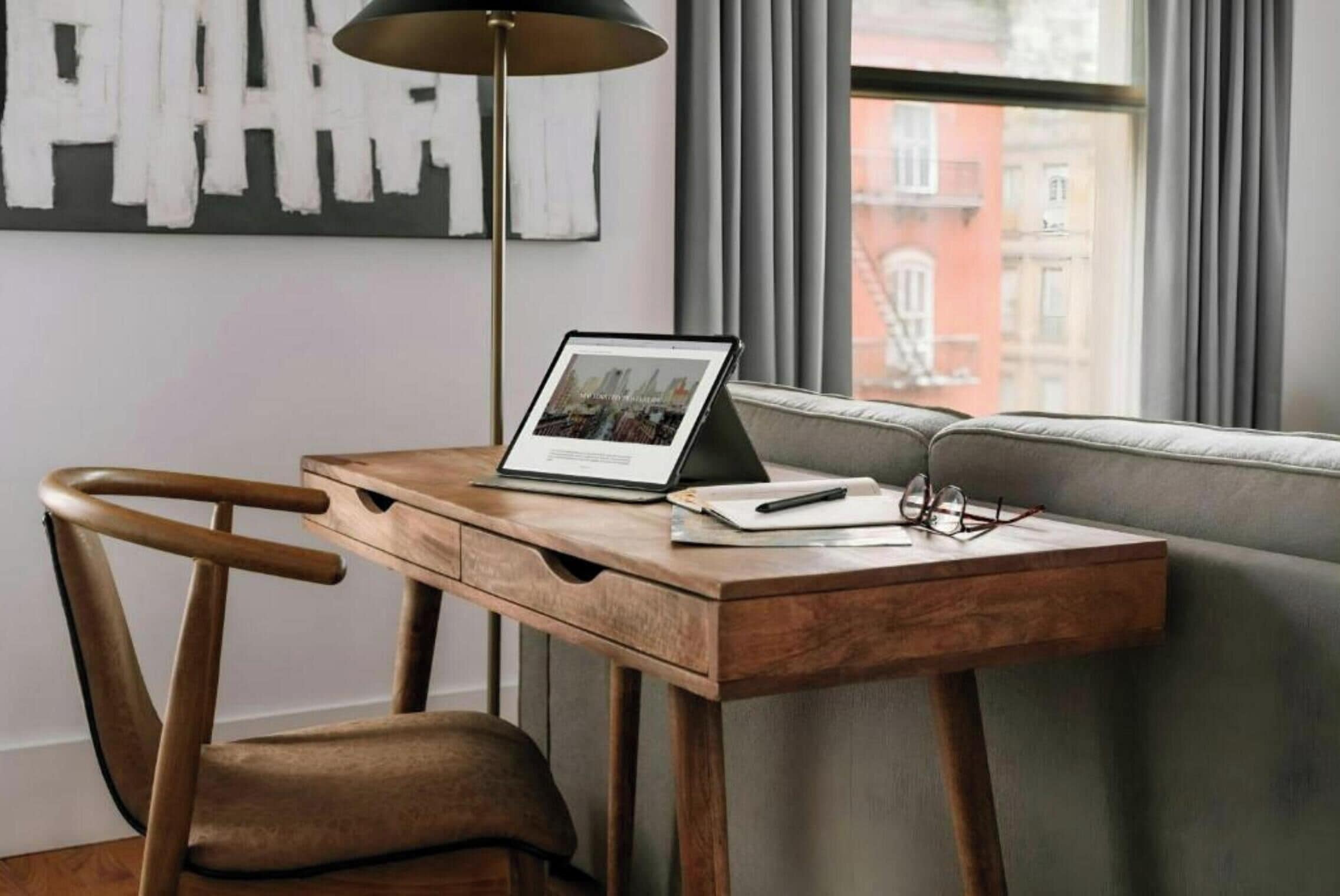 Un escritorio de madera con una computadora portátil, lentes y un cuaderno, ubicado cerca de una ventana con vista a edificios.
