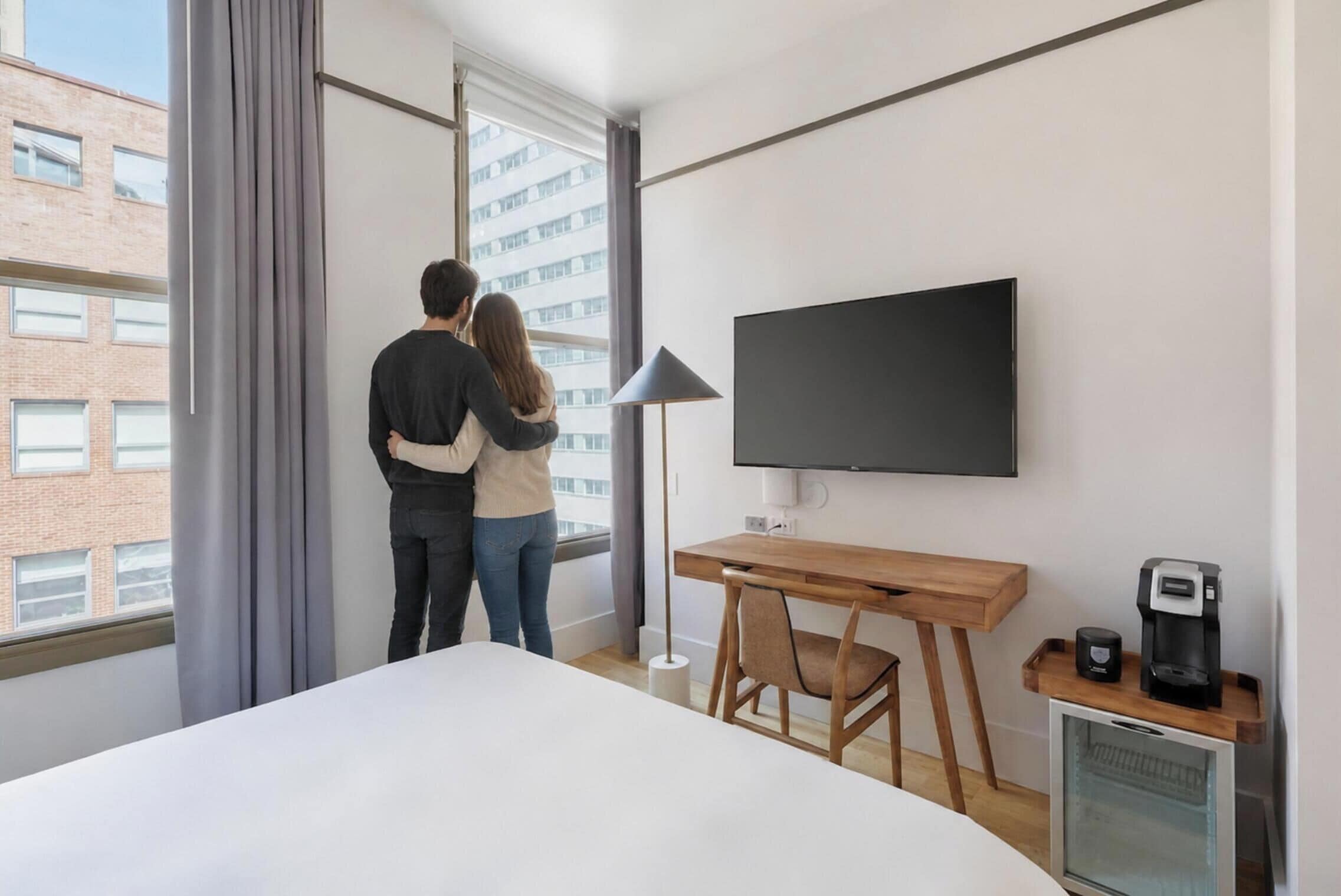 A hotel room with a bed, a desk, a TV, a coffee machine, and a window with curtains.