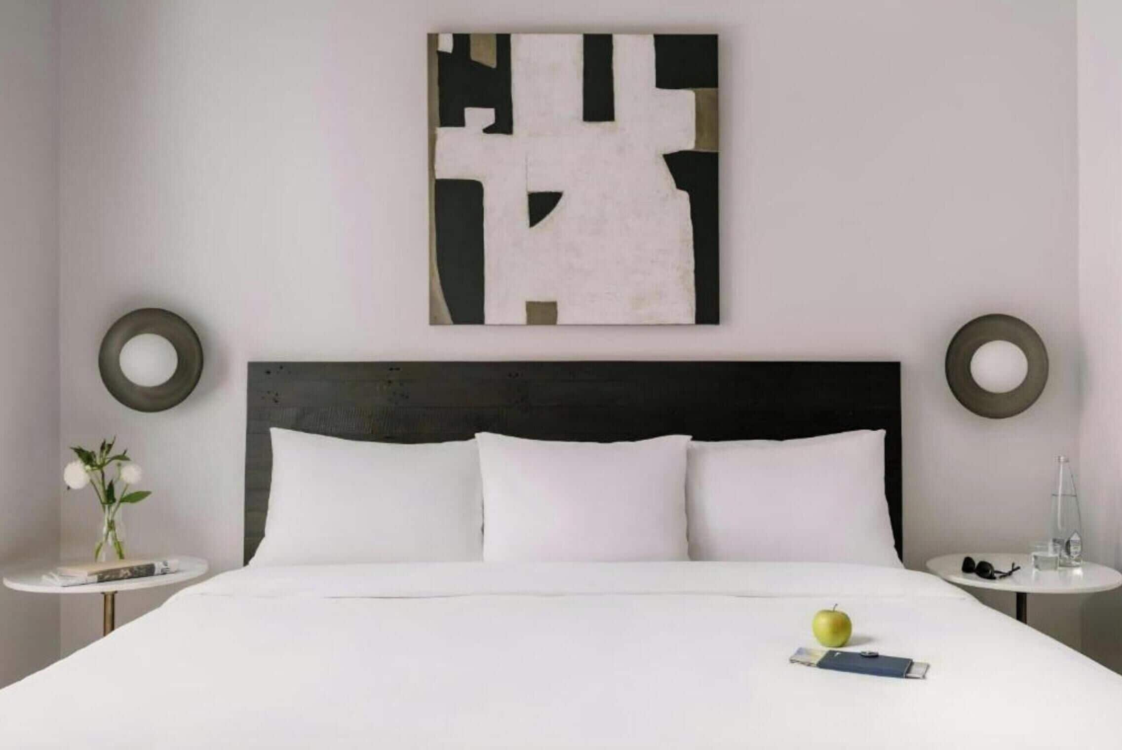 A hotel room with a bed, two bedside tables, a wall art piece, and a round mirror.