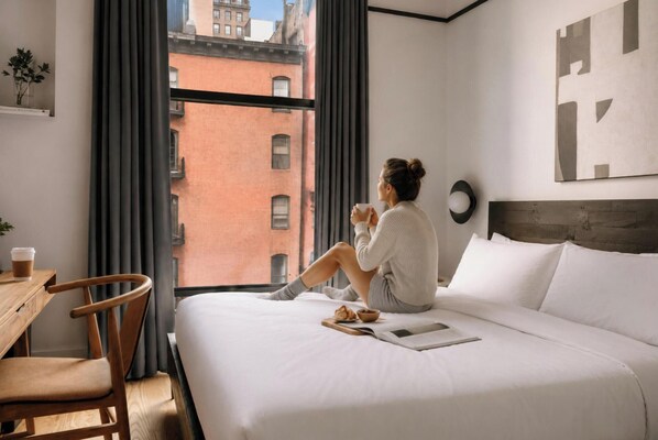 Habitación de hotel con cama, una silla, una mesa, ventana con cortinas y vistas a edificios.