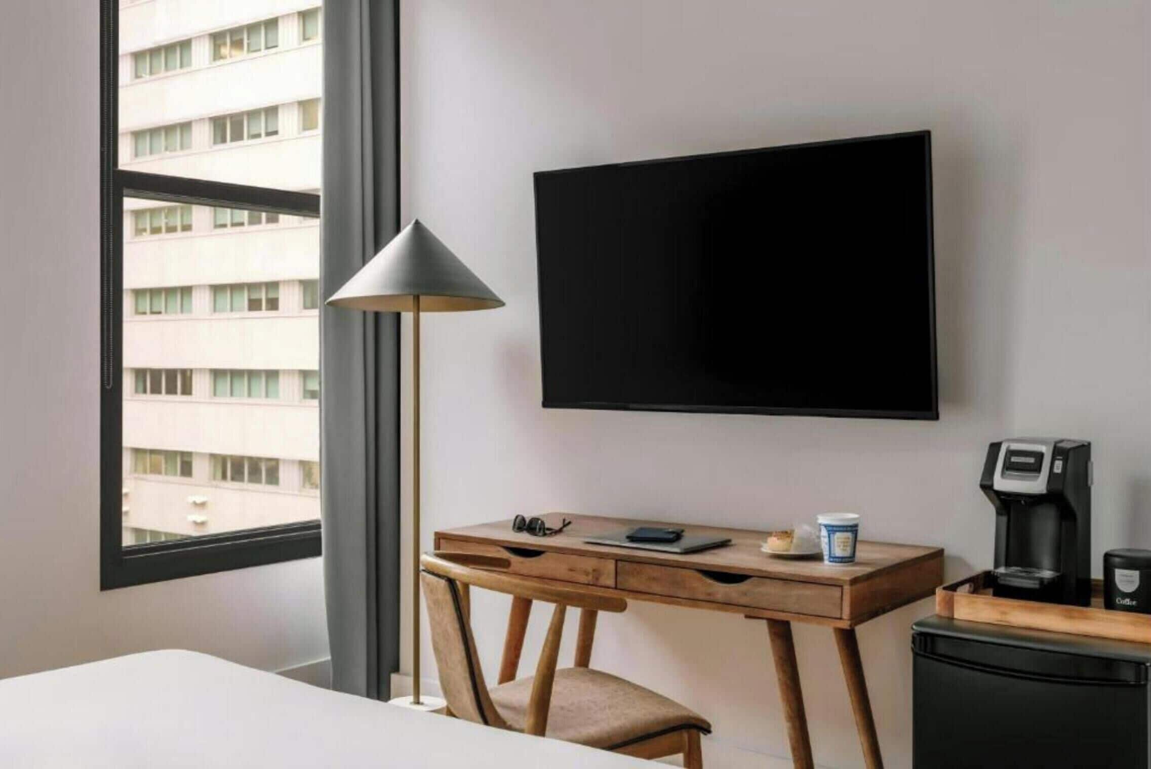 A hotel room with a bed, a desk with a lamp, a coffee machine, and a TV on the wall.