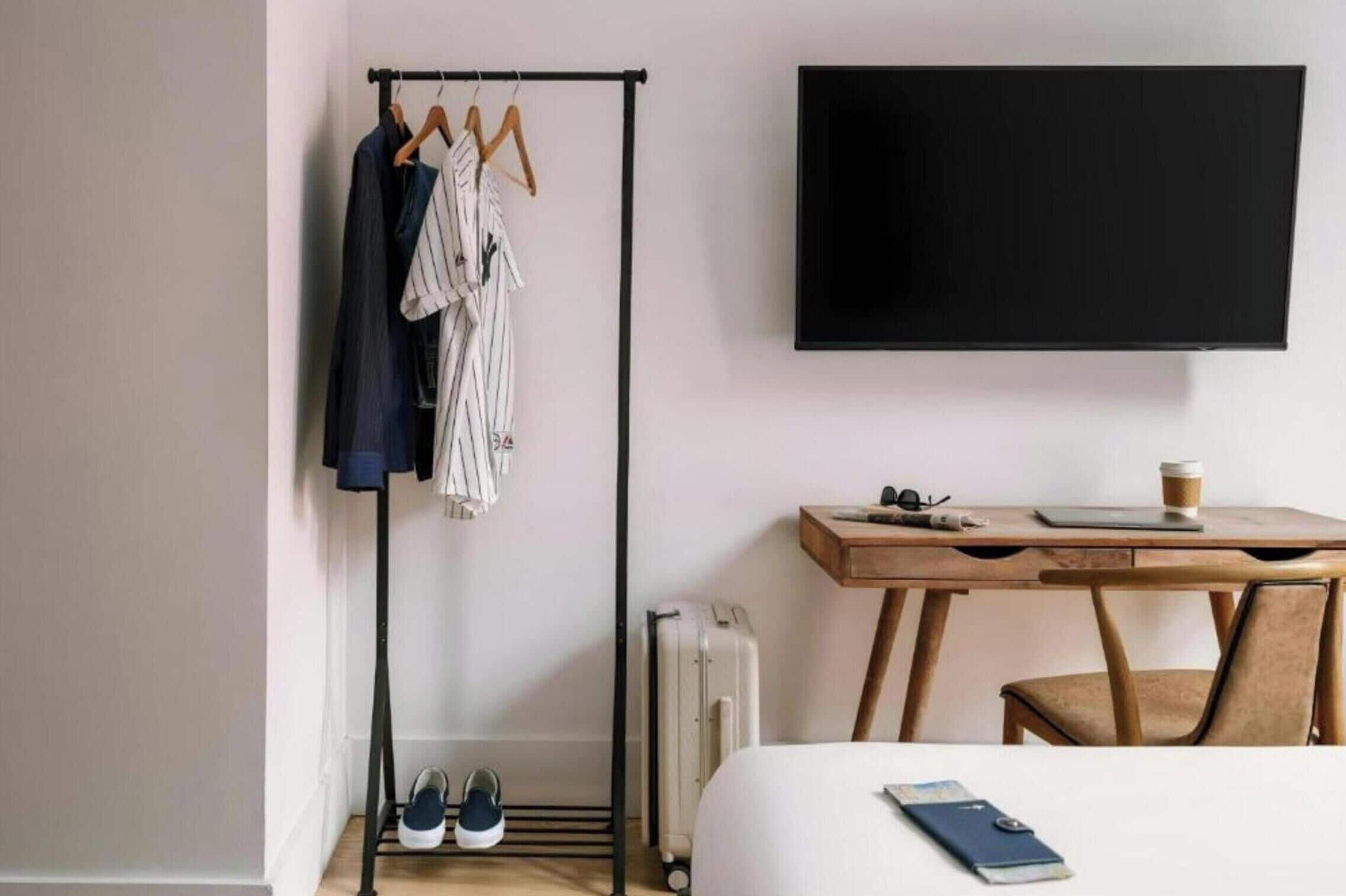 A hotel room with a bed, a desk with a chair, a TV, a coat rack with clothes, a radiator, and a pair of shoes.