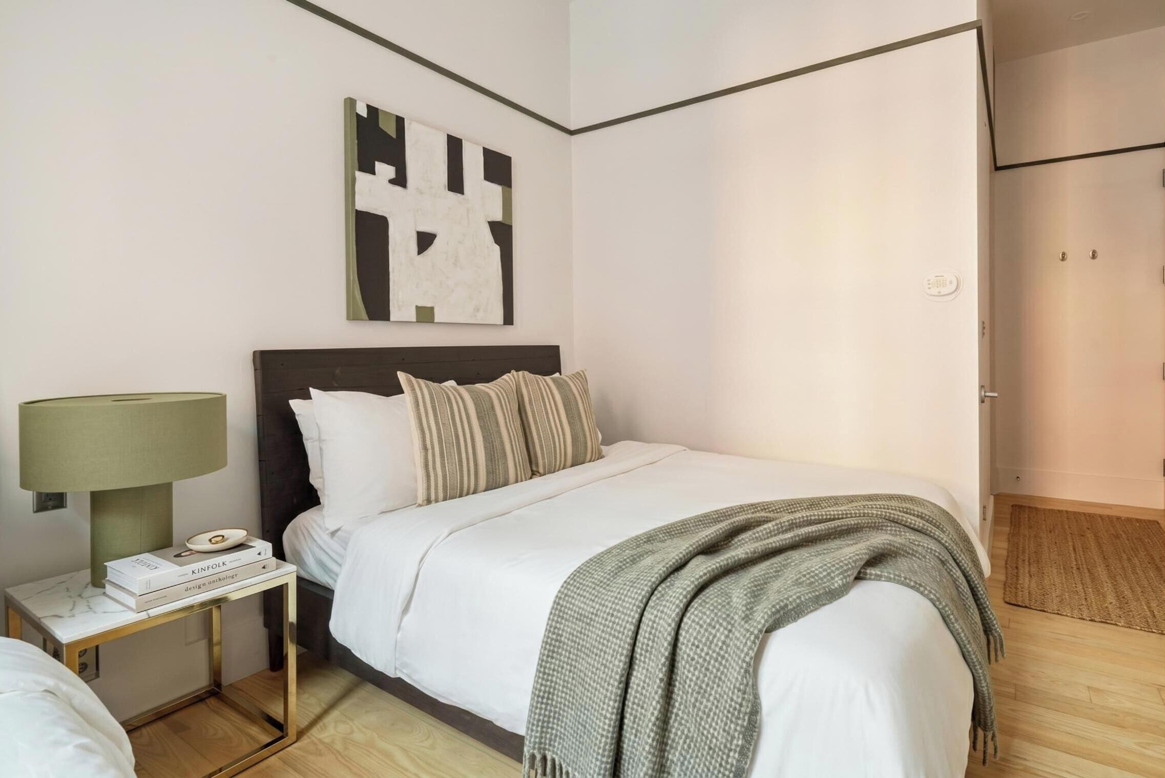 Ein ordentlich eingerichtetes Schlafzimmer mit Bett, Nachttisch, Lampe und abstraktem Kunstwerk an der Wand.