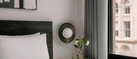 Une chambre d’hôtel avec un lit, un miroir rond, une petite table ornée d’un vase de fleurs et une fenêtre donnant sur un immeuble.