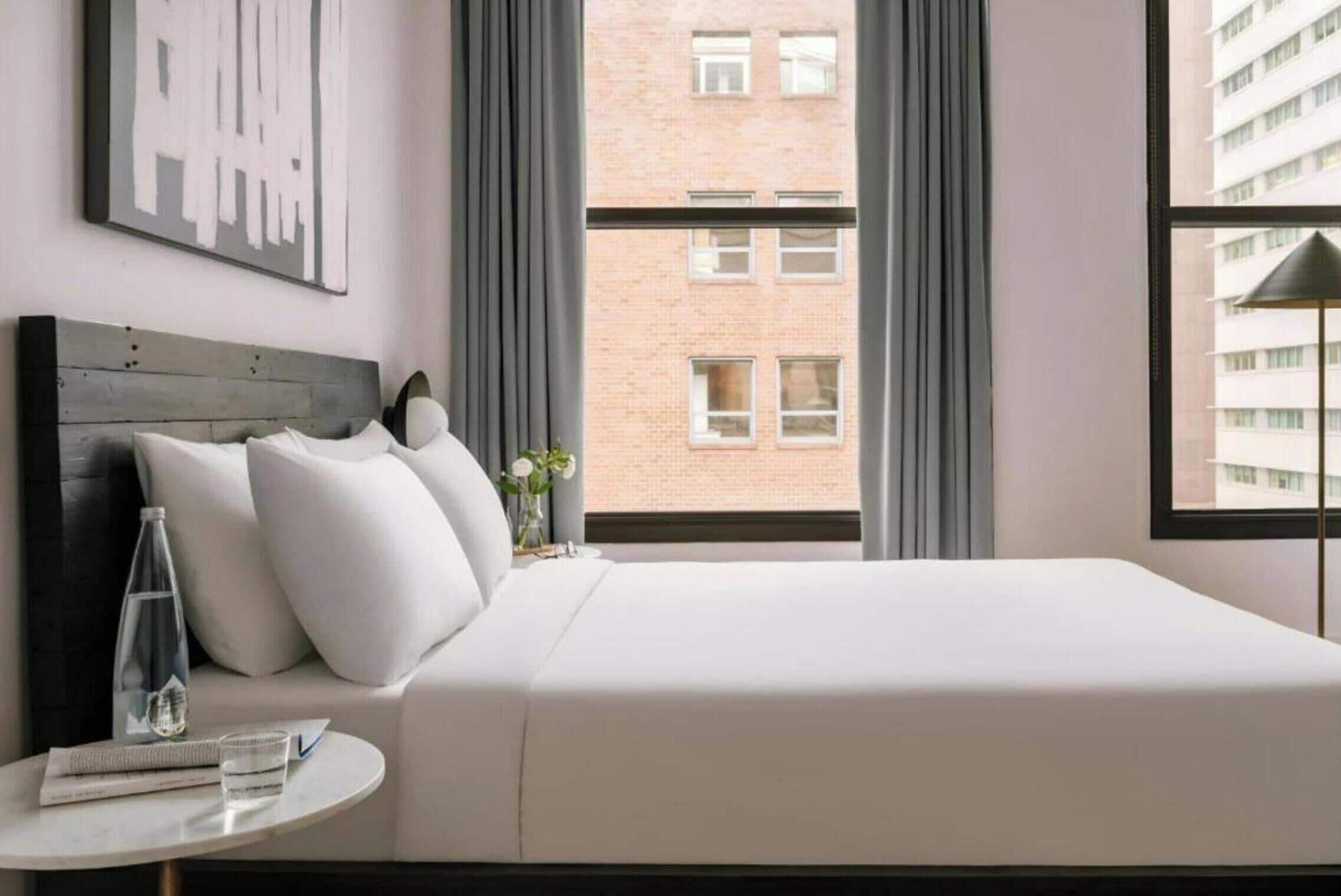 Una habitación de hotel con una cama, una mesa redonda con una botella de vidrio y un libro, y vista a un edificio de ladrillo por la ventana.