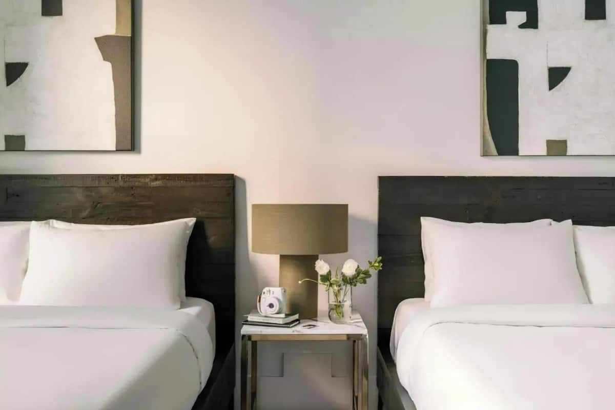 Une chambre d’hôtel avec deux lits, une table de chevet avec une lampe, un appareil photo et un vase avec des fleurs.
