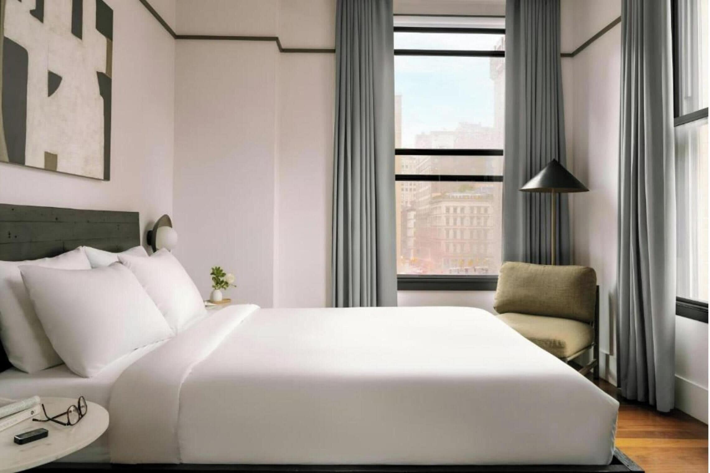 A modern hotel room with a large bed, a chair, a small table, and a view of the city through the window.