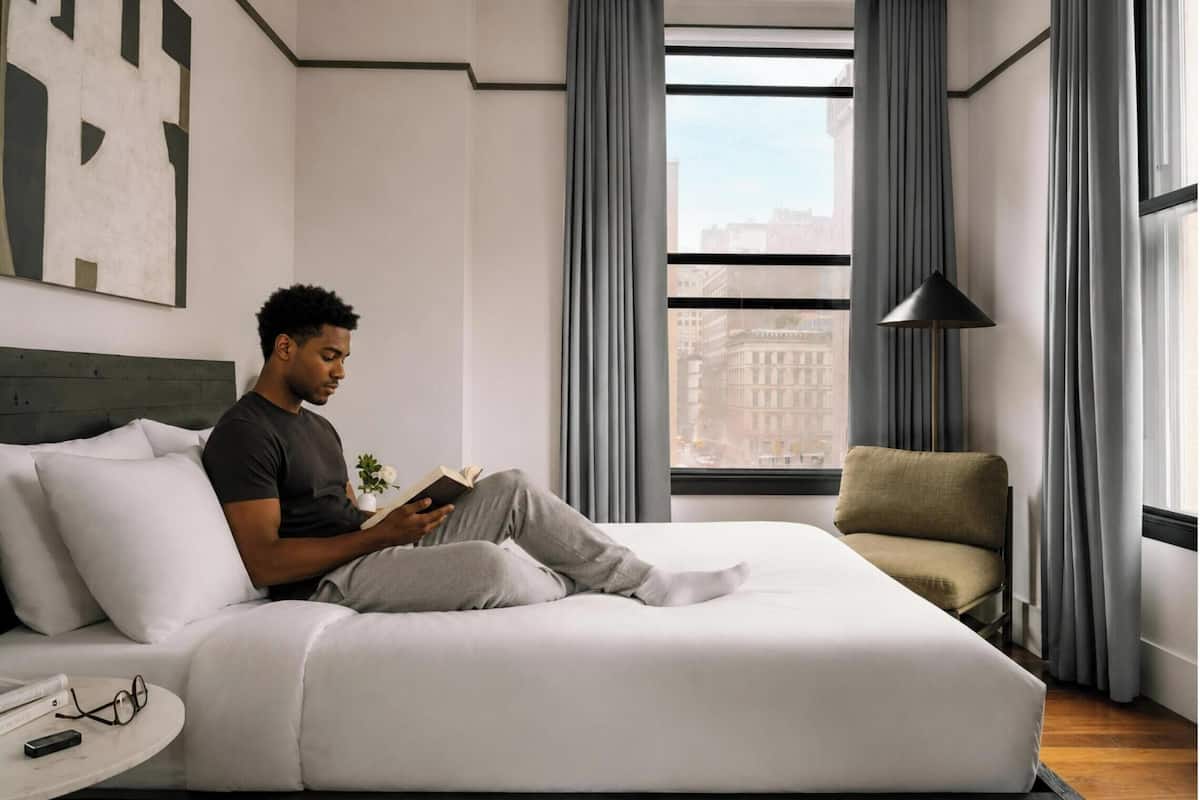 Une chambre d’hôtel moderne, équipée d’un lit, d’un fauteuil de lecture, d’une petite table avec une télécommande et d’une fenêtre avec des rideaux.