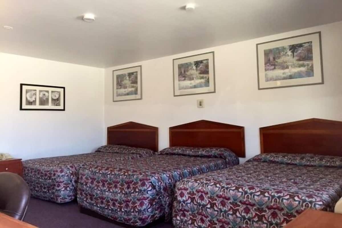 Une chambre d’hôtel avec trois lits simples, une tête de lit en bois, un bureau et une chaise. Trois tableaux encadrés ornent le mur.