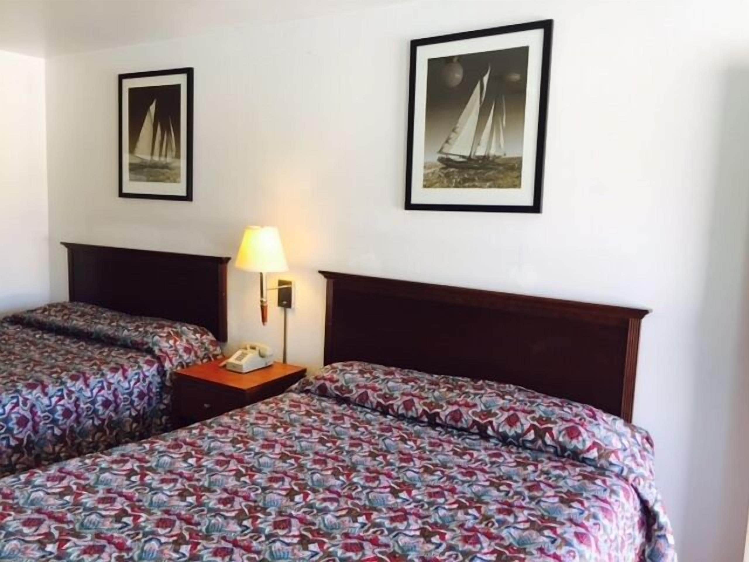 A hotel room with two beds, each with patterned bedding, a bedside table with a lamp, and two framed pictures of sailboats on the wall.