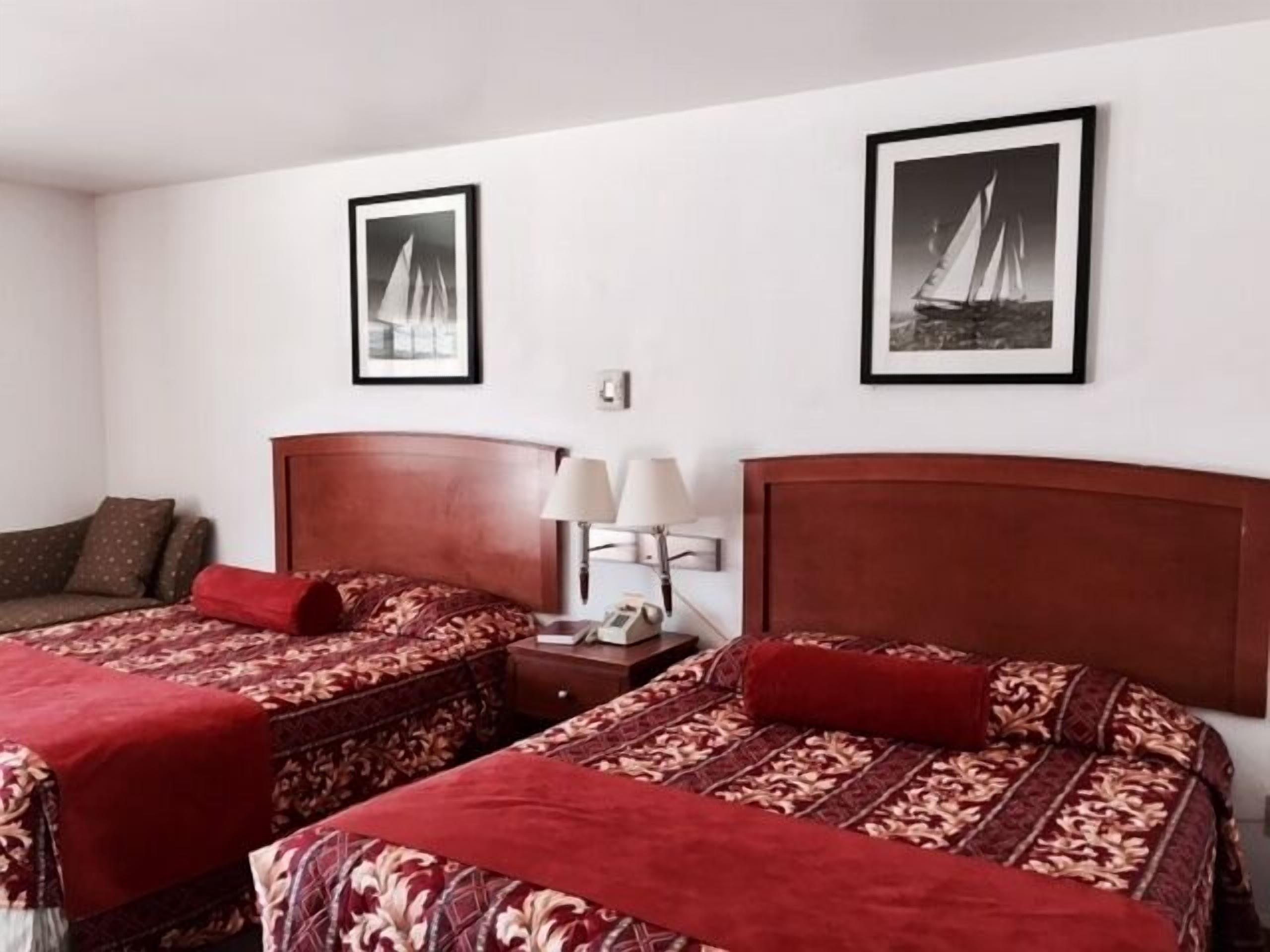 Ein Hotelzimmer mit zwei Betten, jedes mit einer roten, gemusterten Tagesdecke und passenden Kissen. An der Wand befinden sich zwei gerahmte Bilder, jeweils eines über dem Bett. Ein Nachttisch mit Lampe und Telefon steht zwischen den Betten.