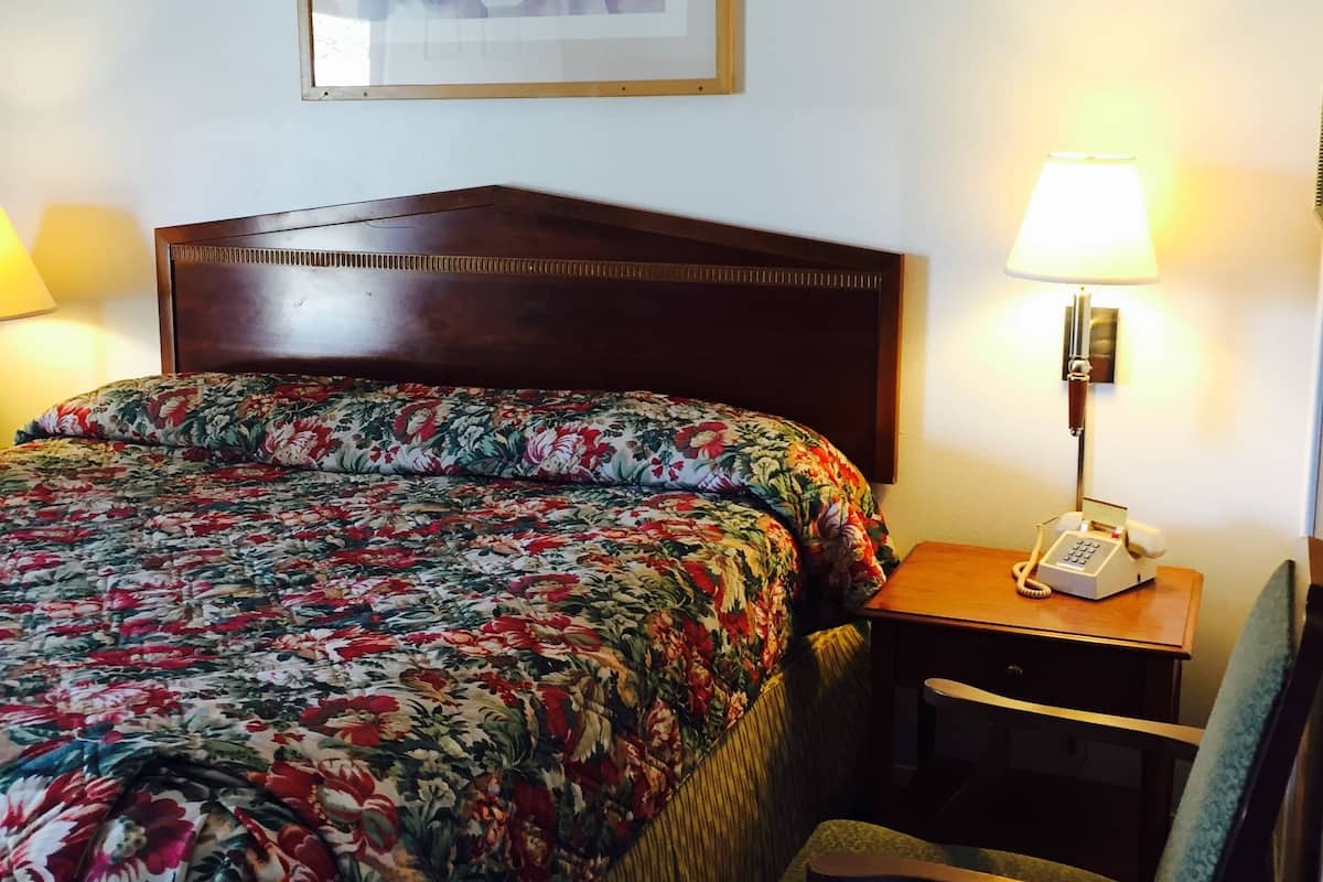 Une chambre d’hôtel avec une couvre-lit à motifs floraux, une tête de lit en bois, un tableau abstrait encadré, une lampe de chevet et un téléphone posé sur une table de chevet en bois.