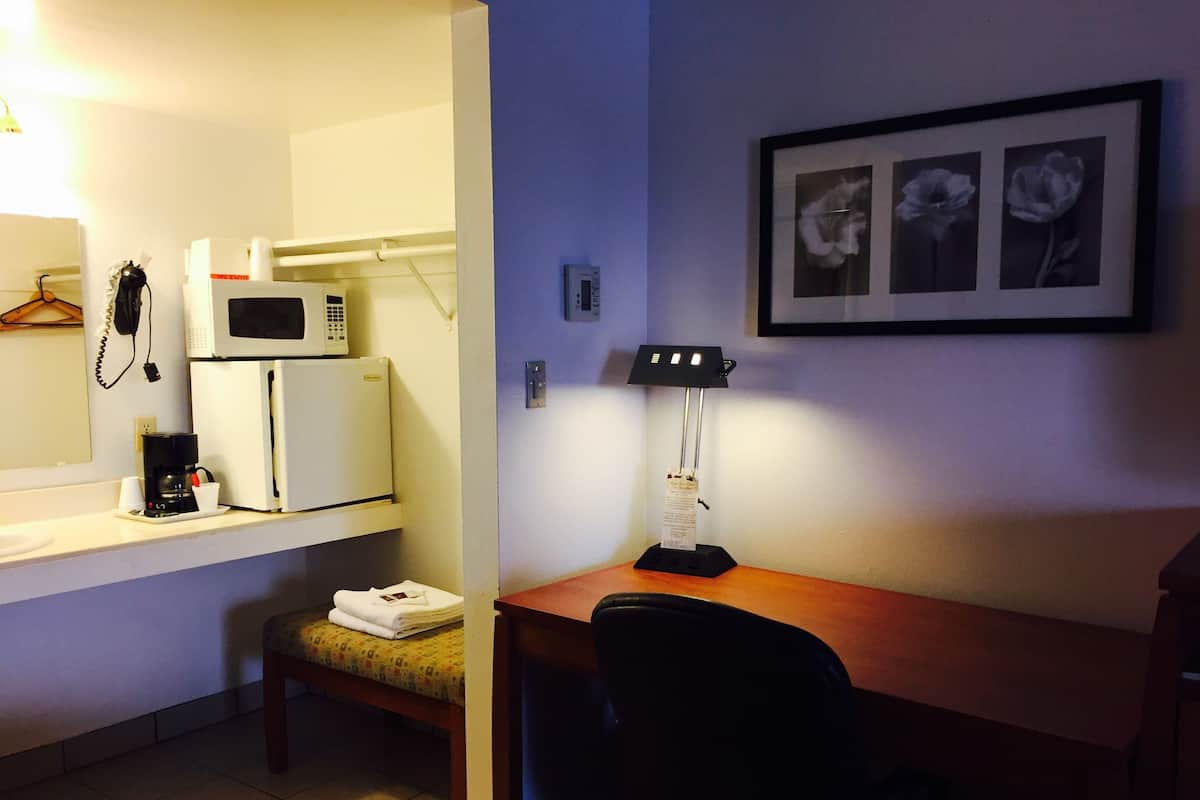Une chambre d’hôtel compacte comprenant un coin cuisine, un bureau avec une chaise, une table de chevet avec une lampe et un tableau encadré au mur.