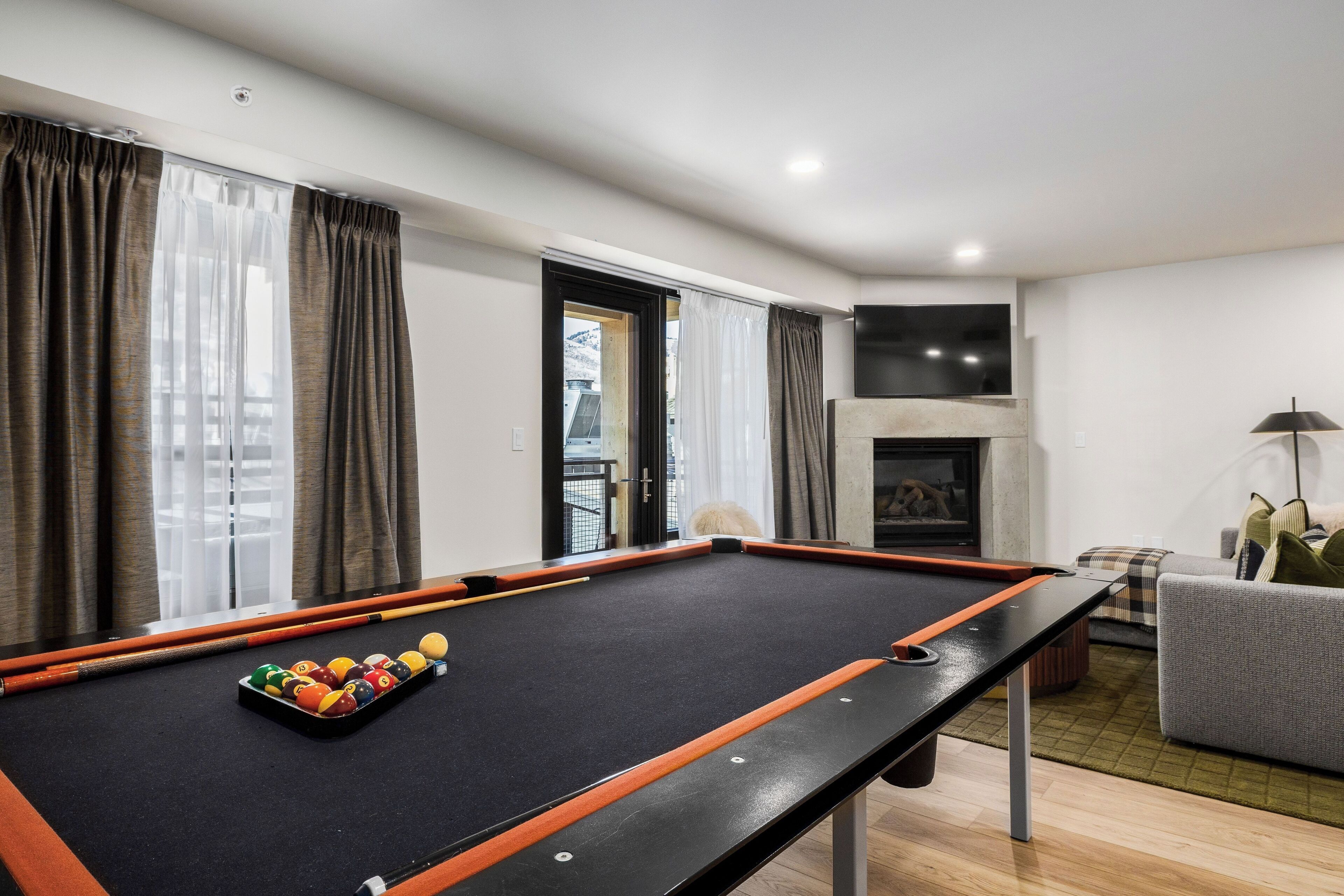A pool table with balls, a sofa, a lamp, and a fireplace in a room with curtains and a view.