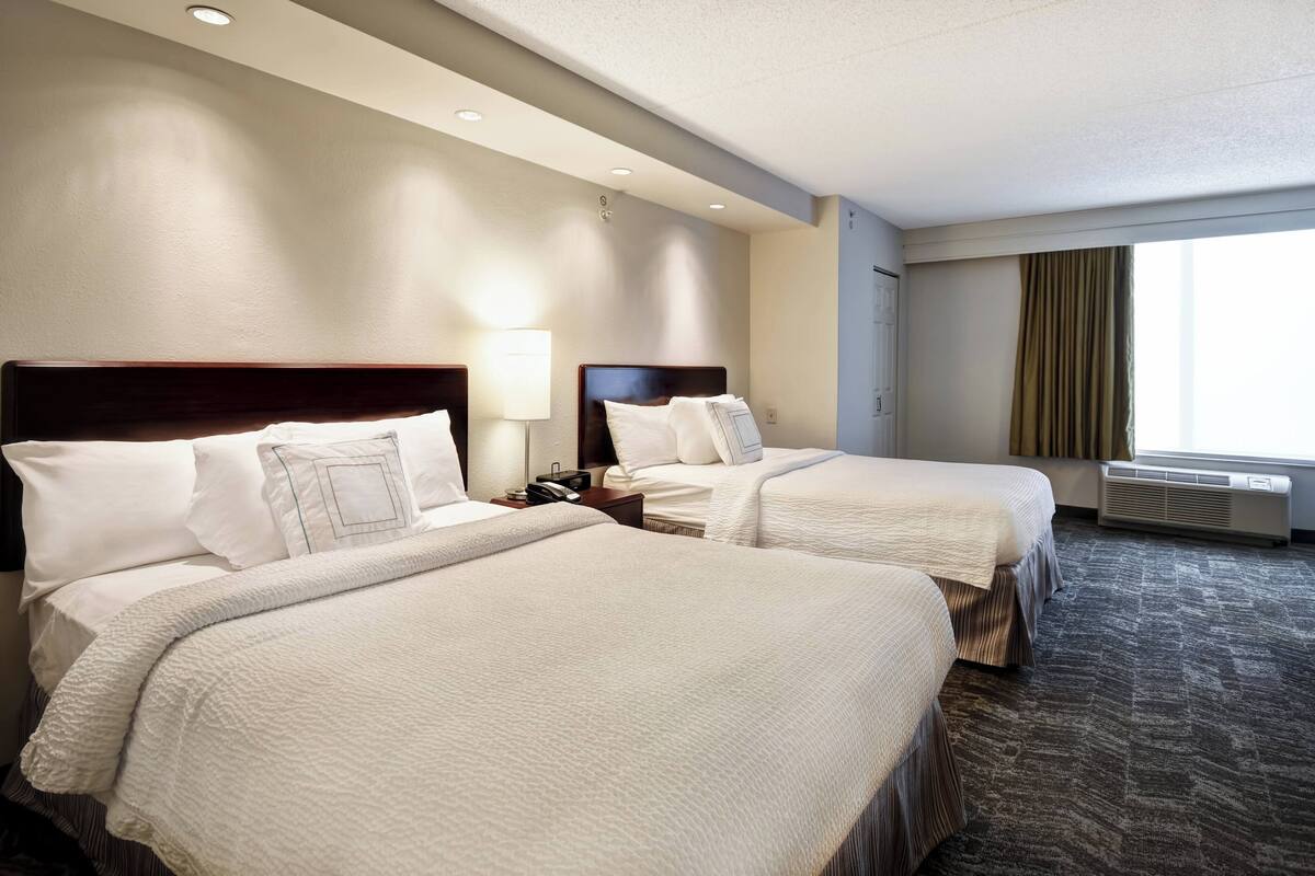 A hotel room with two beds, a nightstand with a lamp, a window with curtains, and a carpeted floor.
