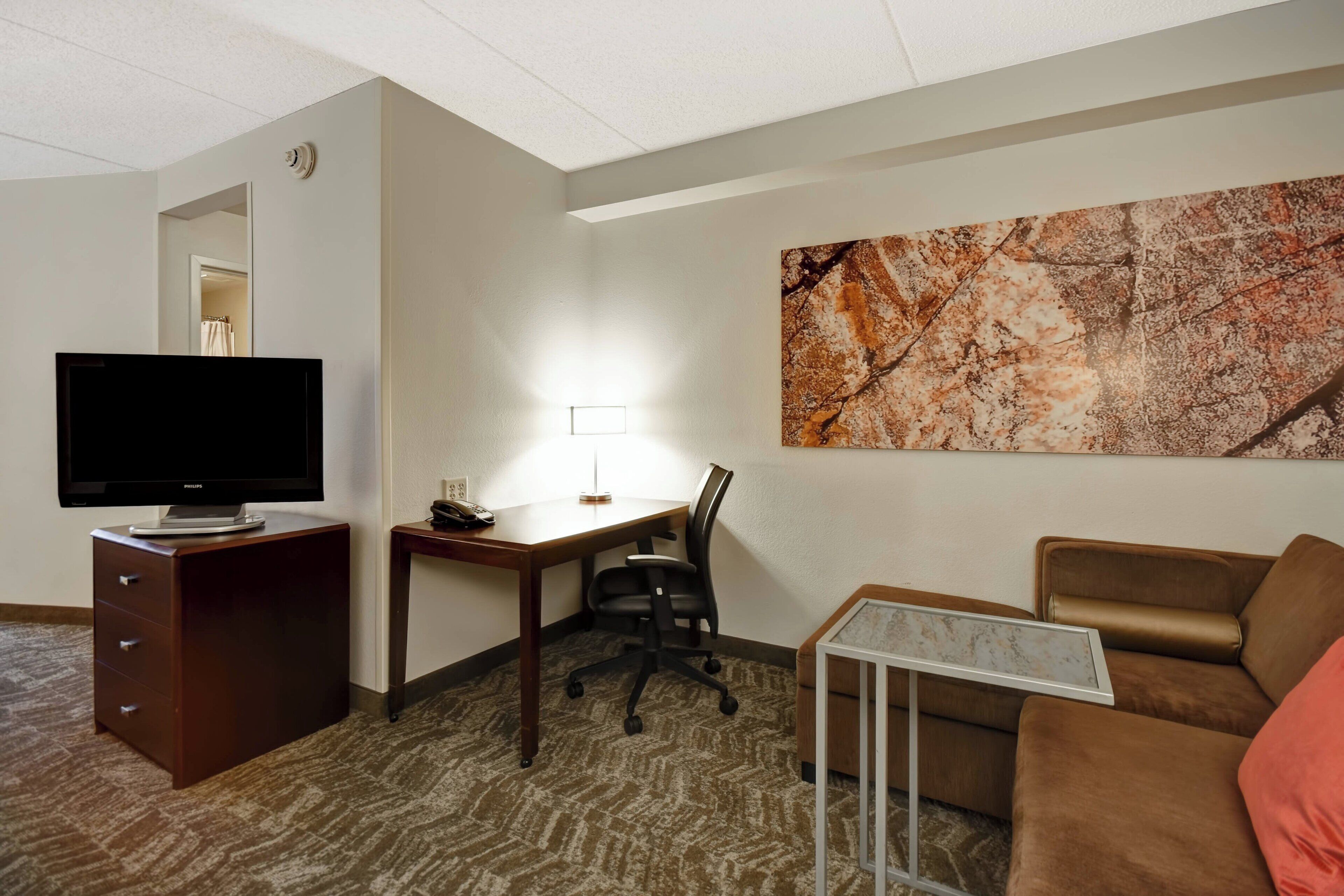 A hotel room with a flat-screen TV, a desk with a lamp, a brown leather sofa, and a marble coffee table.