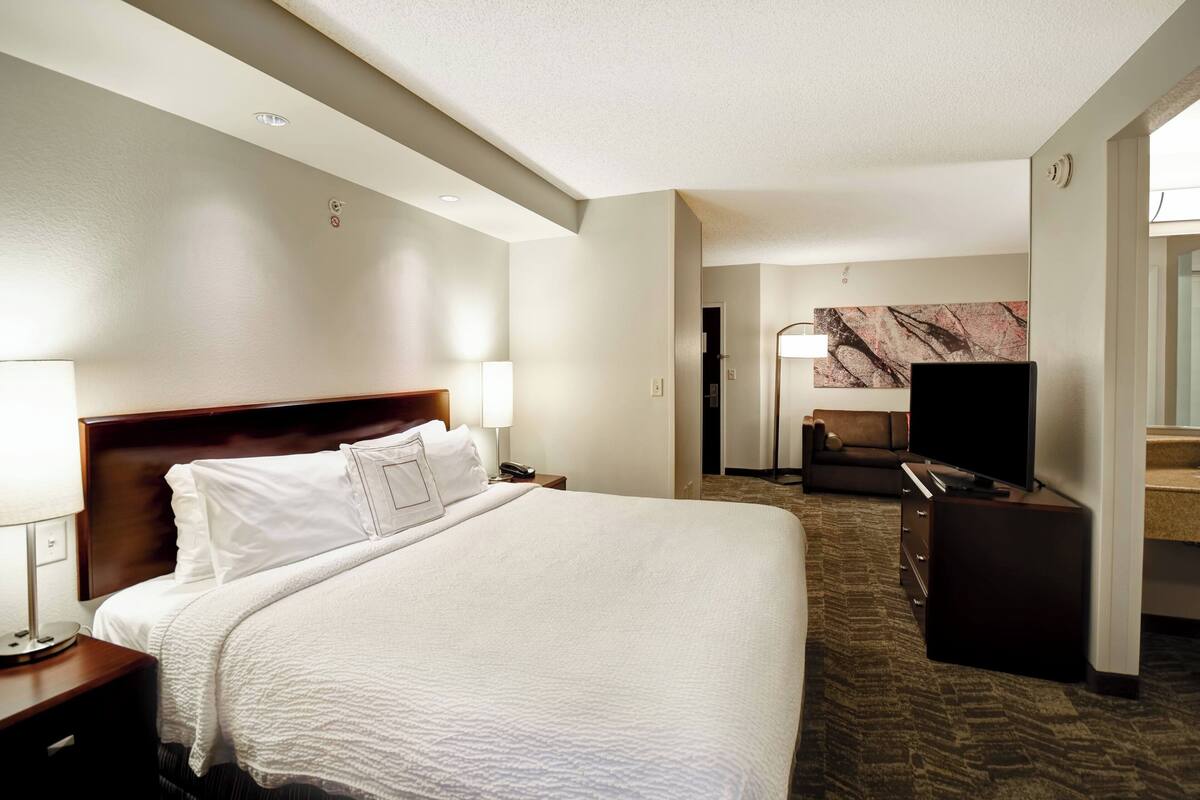 A hotel room with a large bed, a TV, a desk, and a bathroom visible in the background.