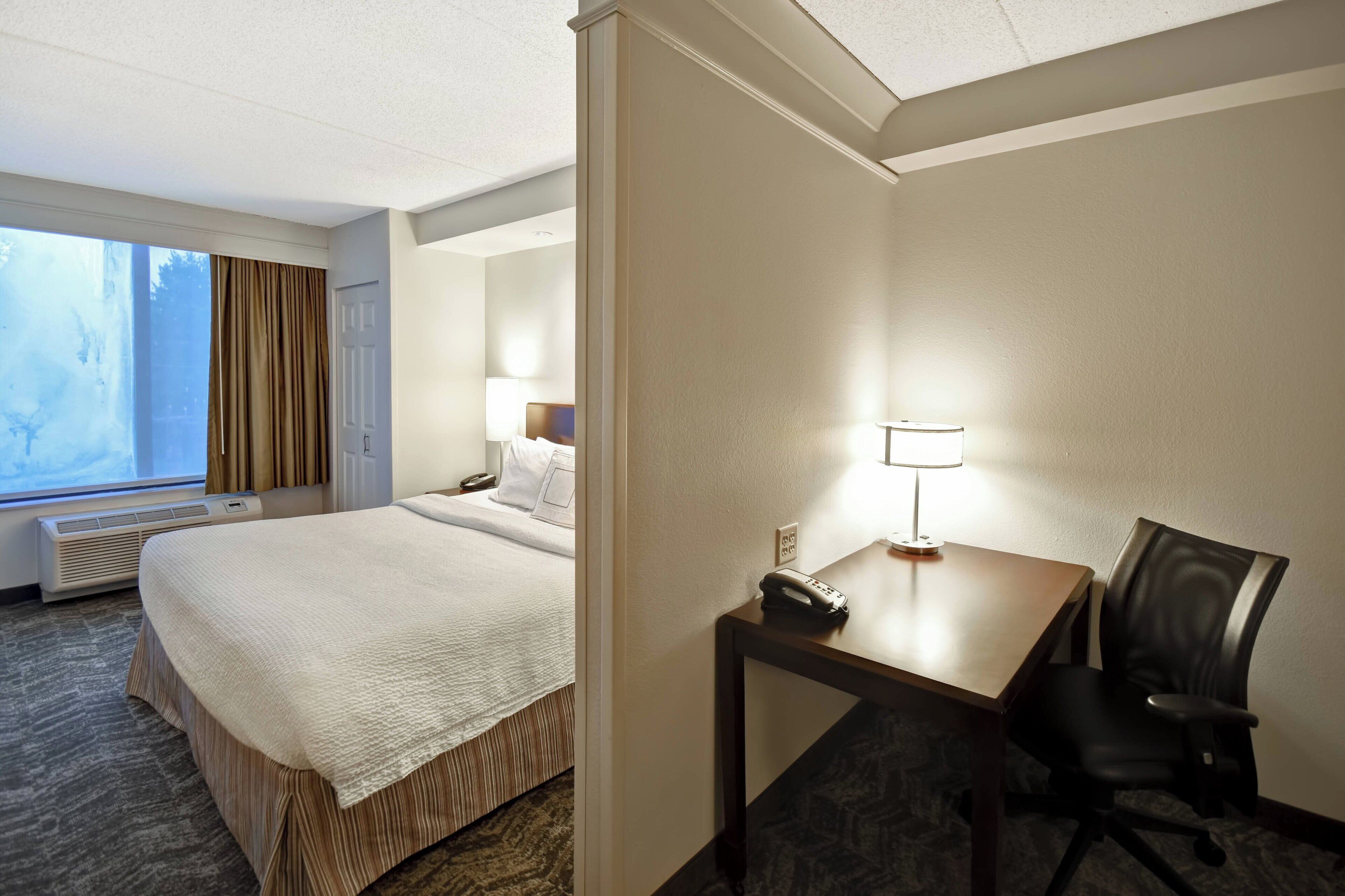 A hotel room with a large bed, a desk with a lamp, a chair, and a window with curtains.