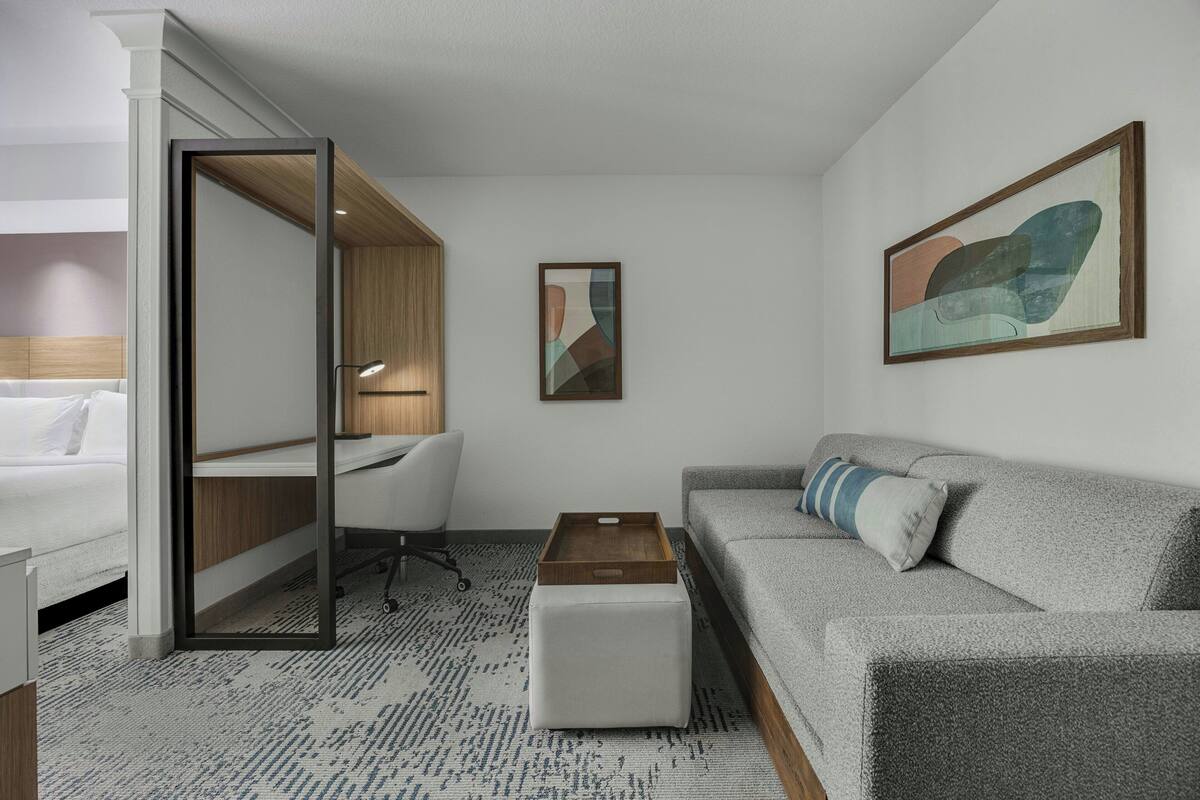 A modern hotel room with a bed, a desk, a gray sofa, a small wooden table, and a painting on the wall.