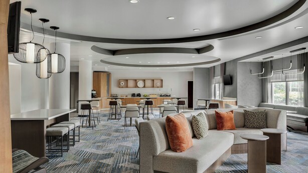 A modern hotel lobby with a curved ceiling, seating area with a sofa and chairs, a bar area, and a kitchen in the background. - SpringHill Suites by Marriott Jacksonville North I-95 Area (Jacksonville)