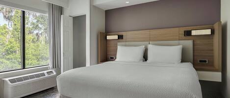 Habitación de hotel moderna con una cama grande, calefacción integrada, ventana con vistas a árboles y cabecero de madera con iluminación incorporada.