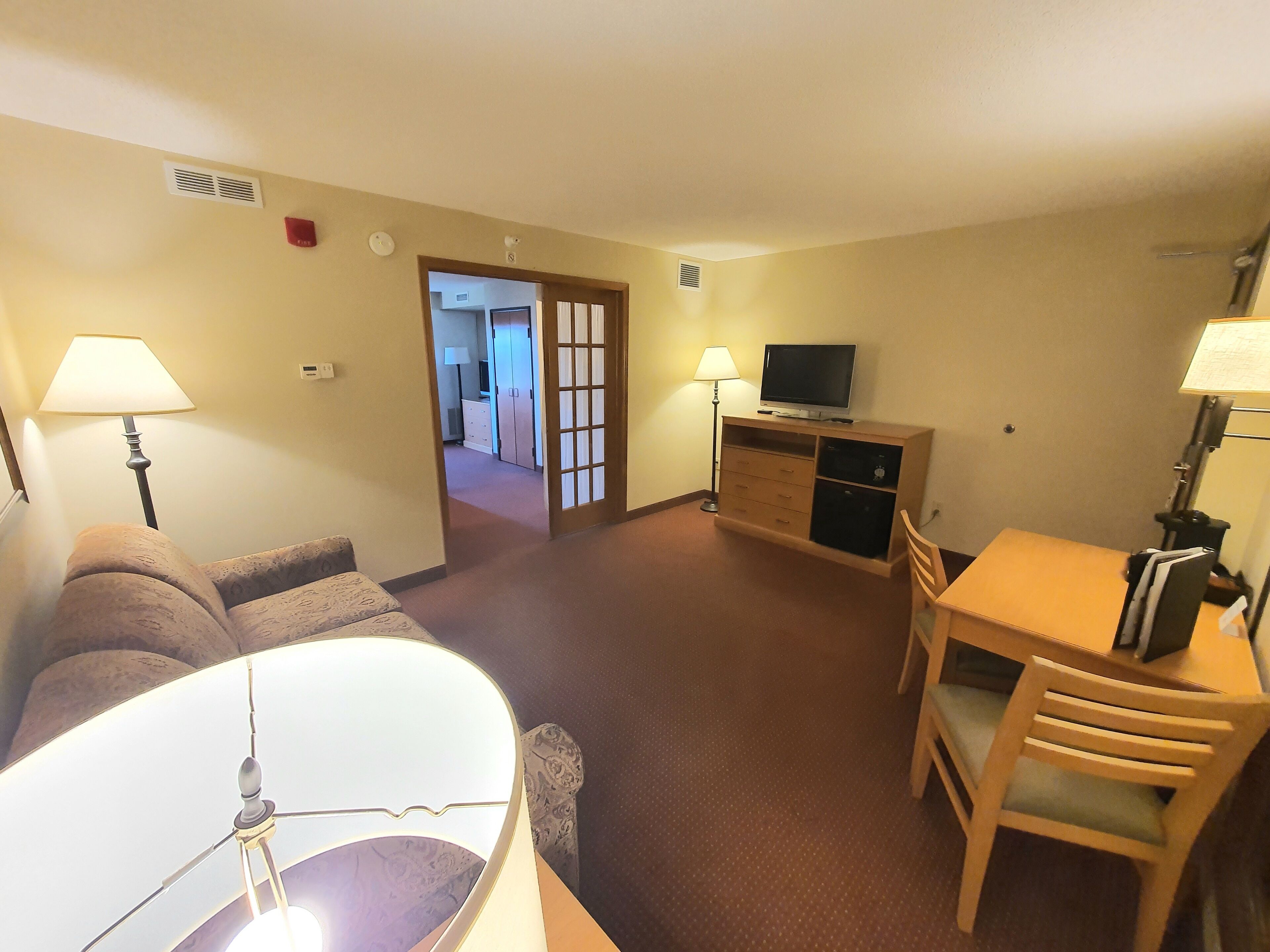 Habitación de hotel con sofá, mesa de comedor con sillas, televisión en un mueble y lámpara.