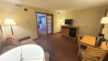 Habitación de hotel con sofá, mesa de comedor con sillas, televisión en un mueble y lámpara.