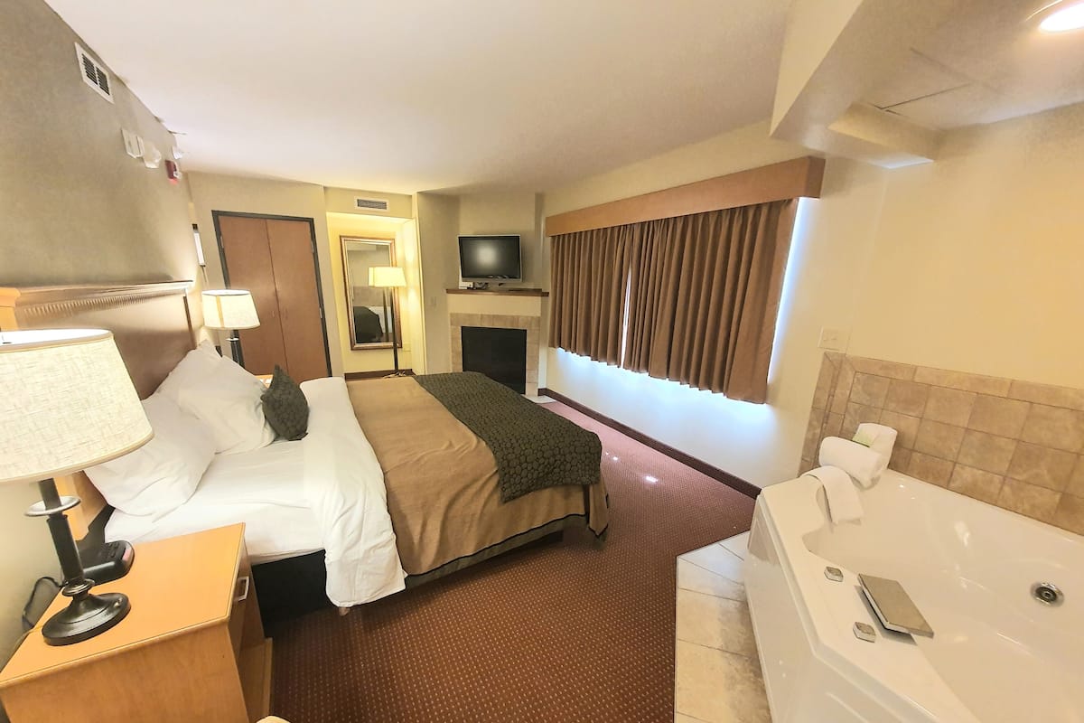 A hotel room with a bed, a desk with a lamp, a TV, a fireplace, a large window with curtains, and a bathtub.
