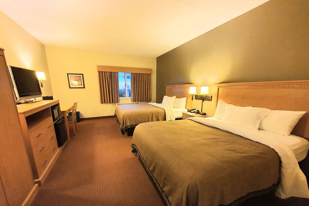 A hotel room with two beds, a desk with a television, a window with curtains, and a nightstand with a lamp.