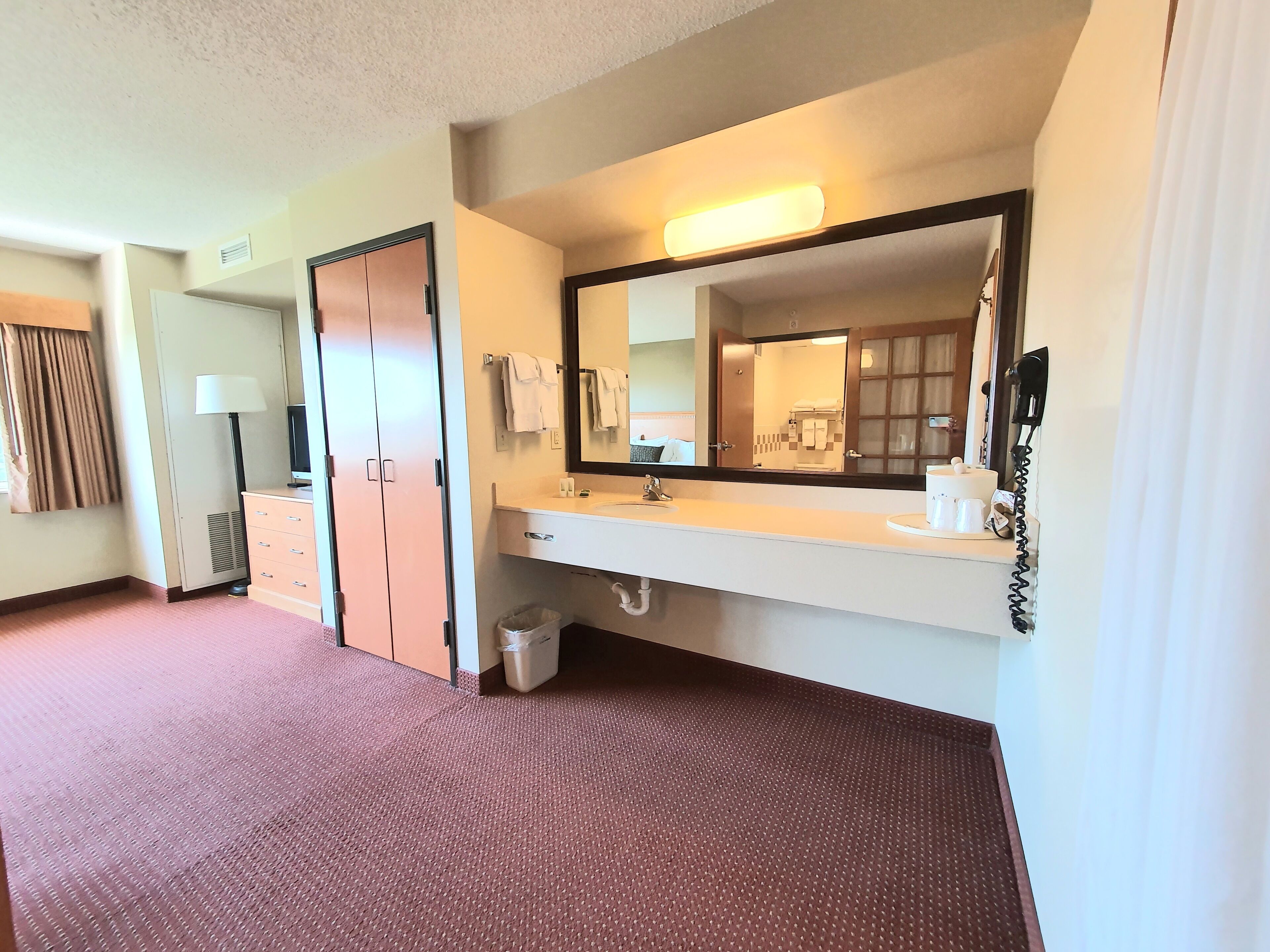 Habitación de hotel con un espejo grande, una tocador con lavamanos, un cubo de basura y un armario.