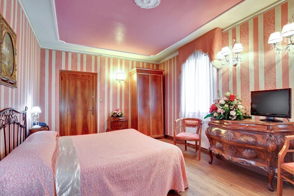 Una habitación de hotel con papel tapiz a rayas, una cama con colcha rosa, un aparador de madera, un televisor y una silla.