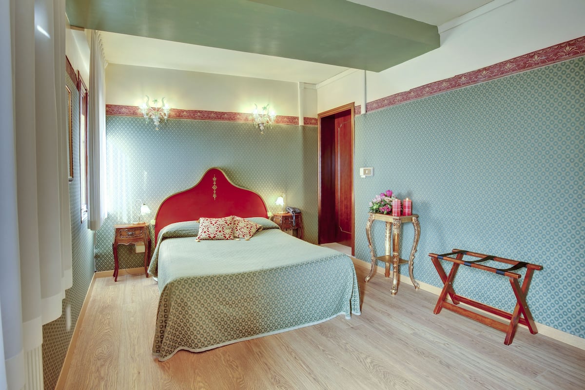 Un dormitorio con cabecera roja, cama con ropa de cama estampada, mesita de noche con lámpara, una pequeña mesa con un jarrón de flores y una silla de madera.