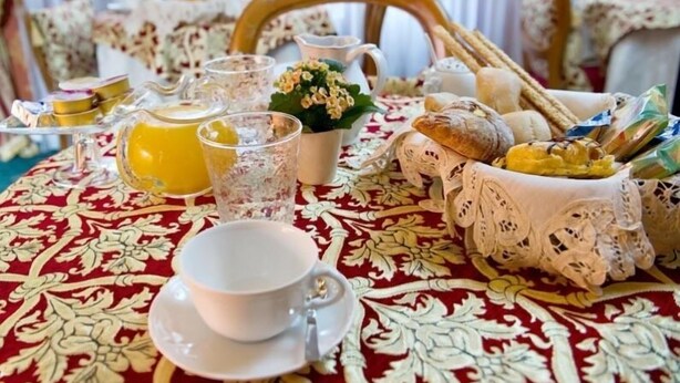 Una mesa vestida con un mantel rojo estampado, una taza y platillo blancos, una jarrón, vasos, y una cesta con pan y pasteles.