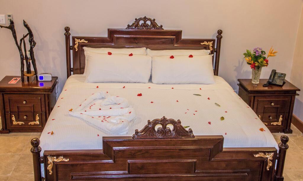 Ein großes, verziertes Bett mit weißen Leinen und Rosenblättern, die auf der Bettdecke verstreut sind. Zwei Nachttische, einer mit einer Lampe und einem Blumenstrauß.