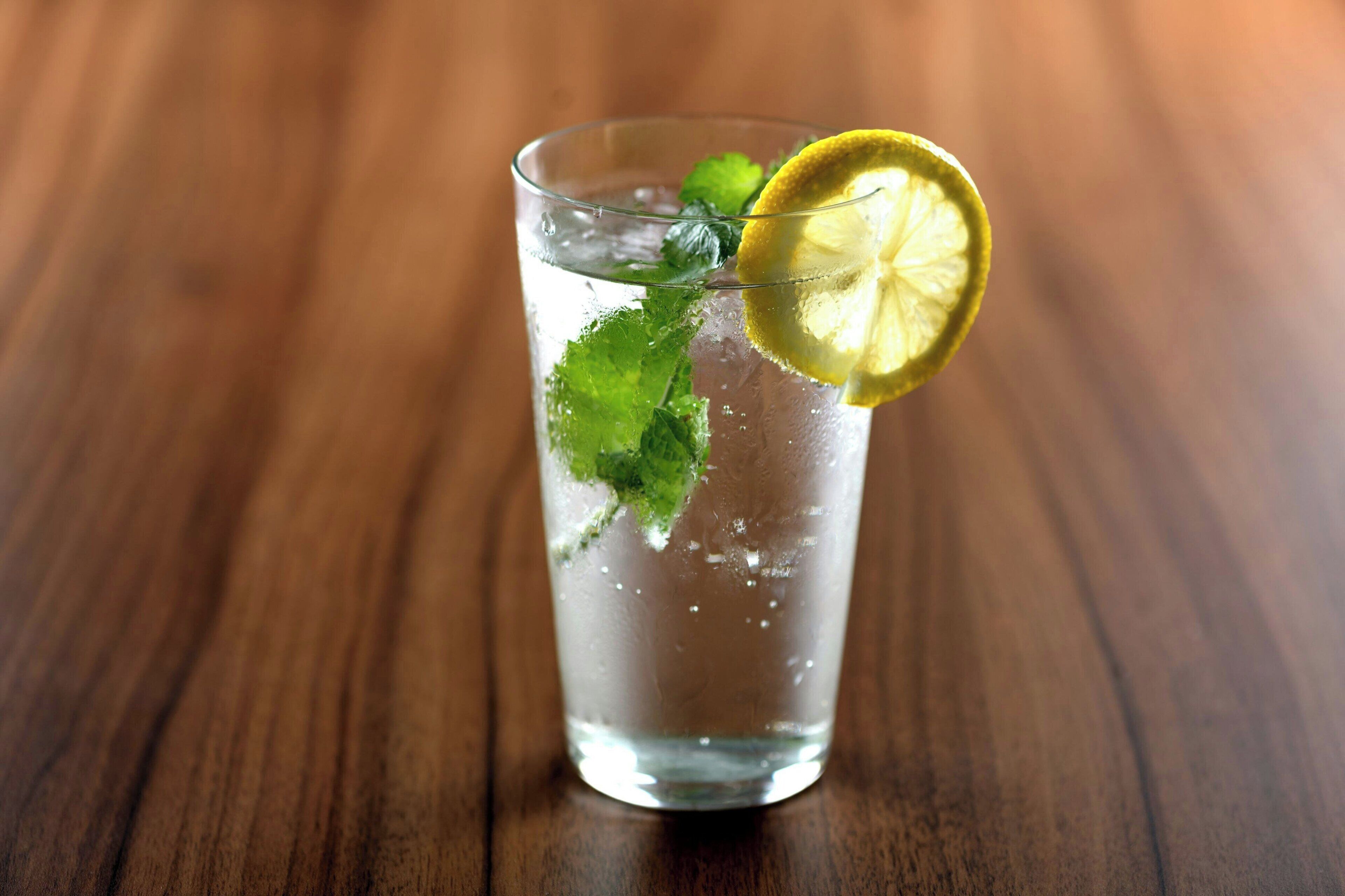 Un verre transparent rempli de glaçons, de feuilles de menthe et d’une tranche de citron, posé sur une surface en bois.