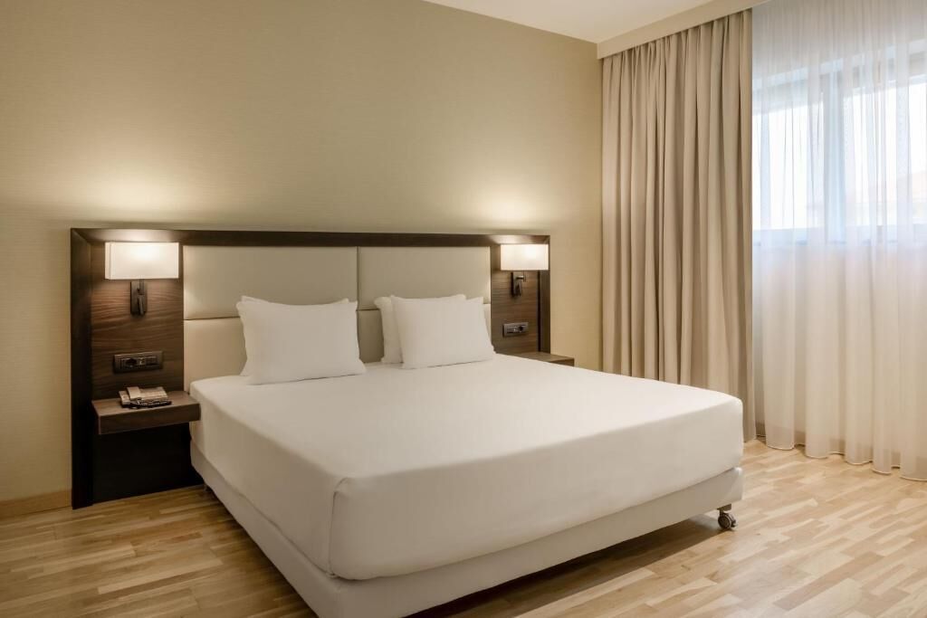 A hotel room with a large bed, two bedside lamps, a nightstand, and a window with curtains.