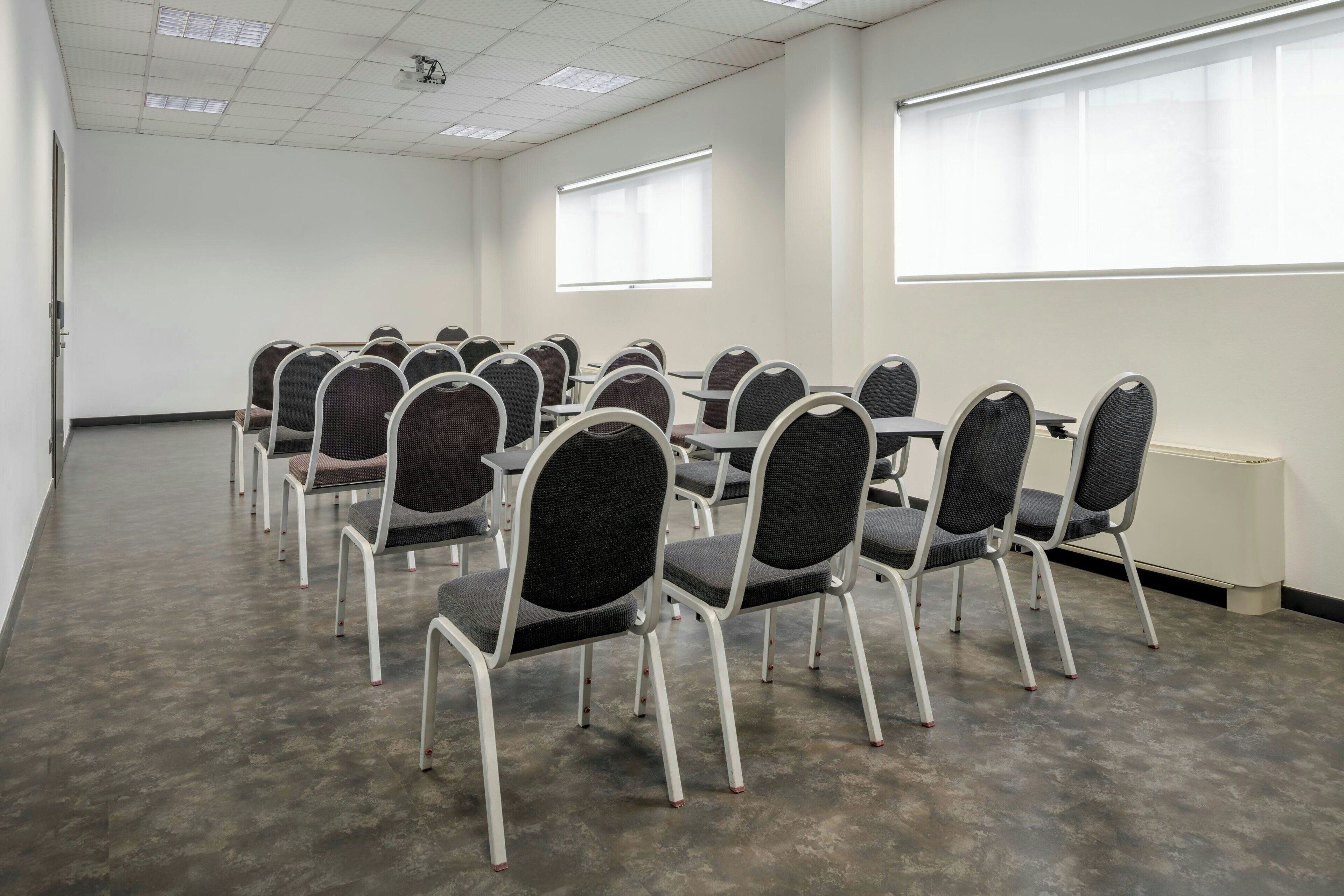 Un salón de conferencias con filas de sillas y mesas, paredes blancas y ventanas amplias.