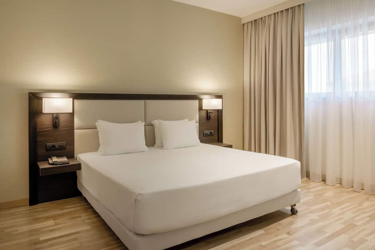 Ein Hotelzimmer mit einem großen Bett, zwei Nachttischen, einer Ablagefläche und einer Wandlampe, sowie einem Fenster mit Vorhängen.