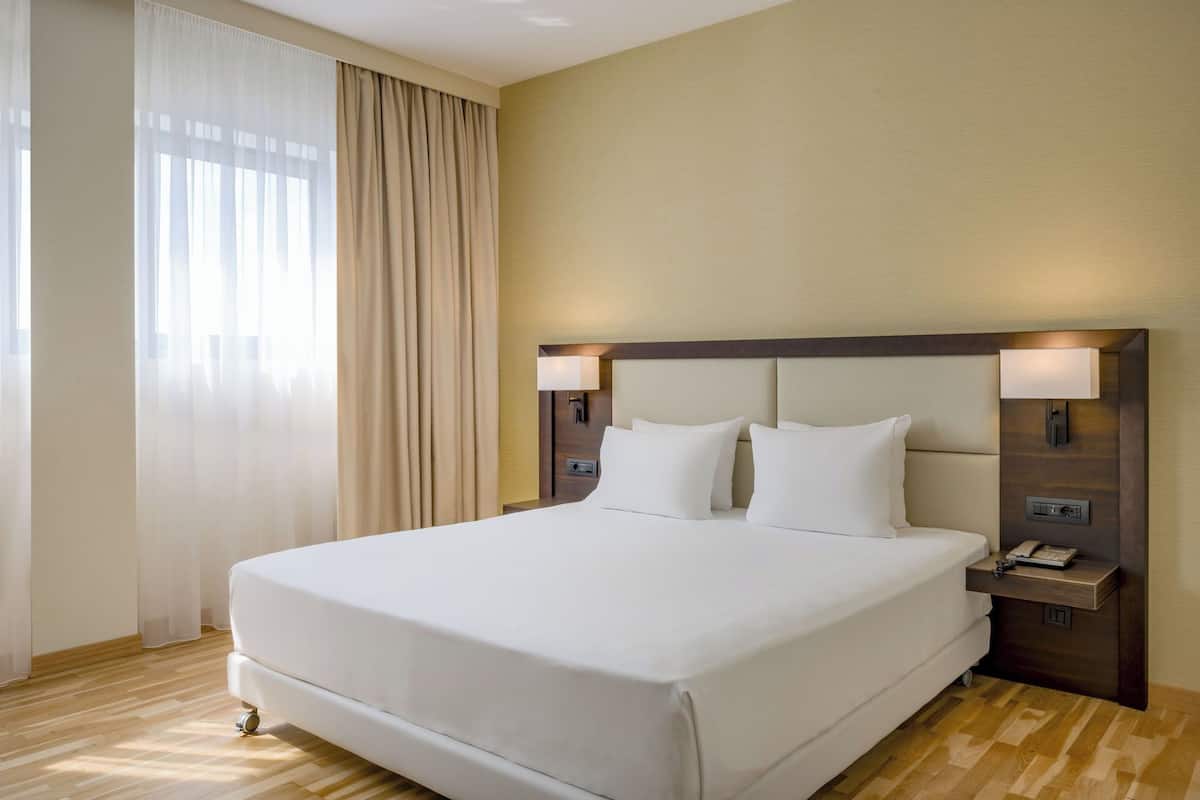 Ein Hotelzimmer mit einem großen Bett, zwei Nachttischen mit Lampen, Holzboden und einem Fenster mit leichtem Vorhang.