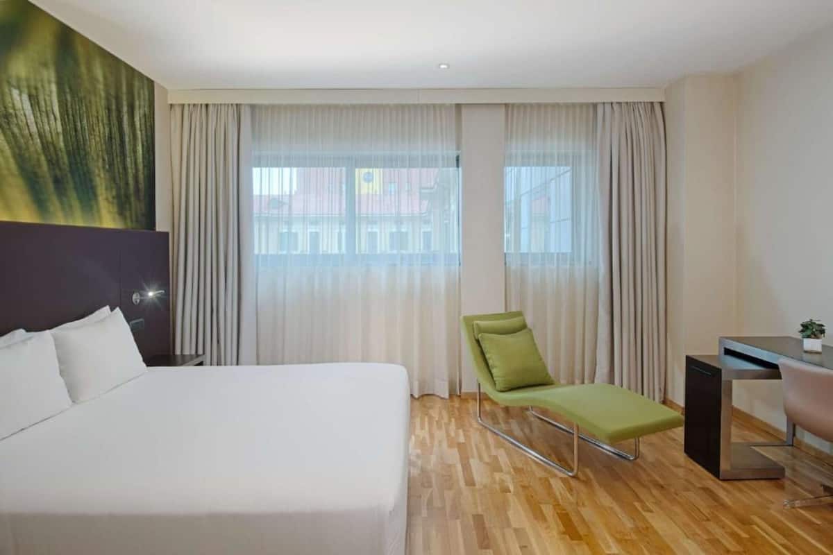 Ein Hotelzimmer mit einem großen Bett, einem Schreibtisch, einem Sessel und Blick durch durchscheinende Vorhänge nach draußen.