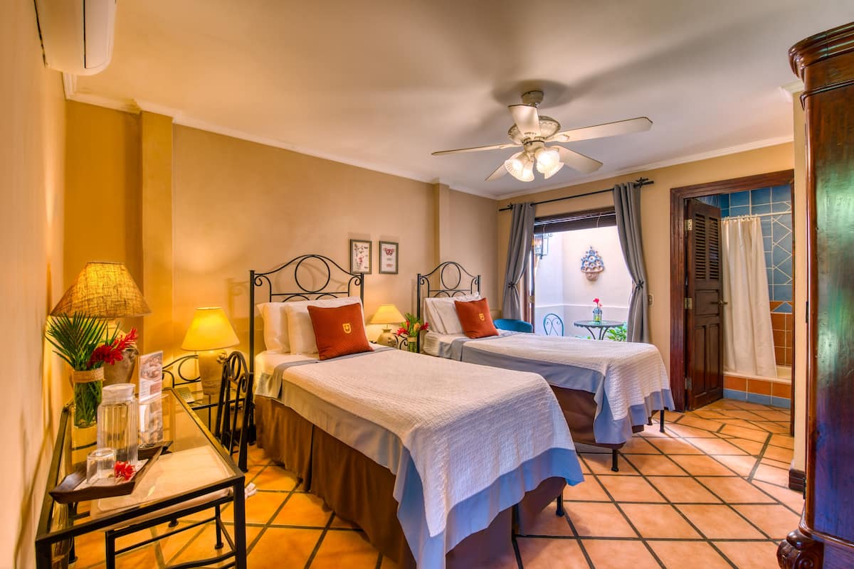 A hotel room with two beds, a ceiling fan, a window with curtains, a small table with a lamp, and a view of the outside.